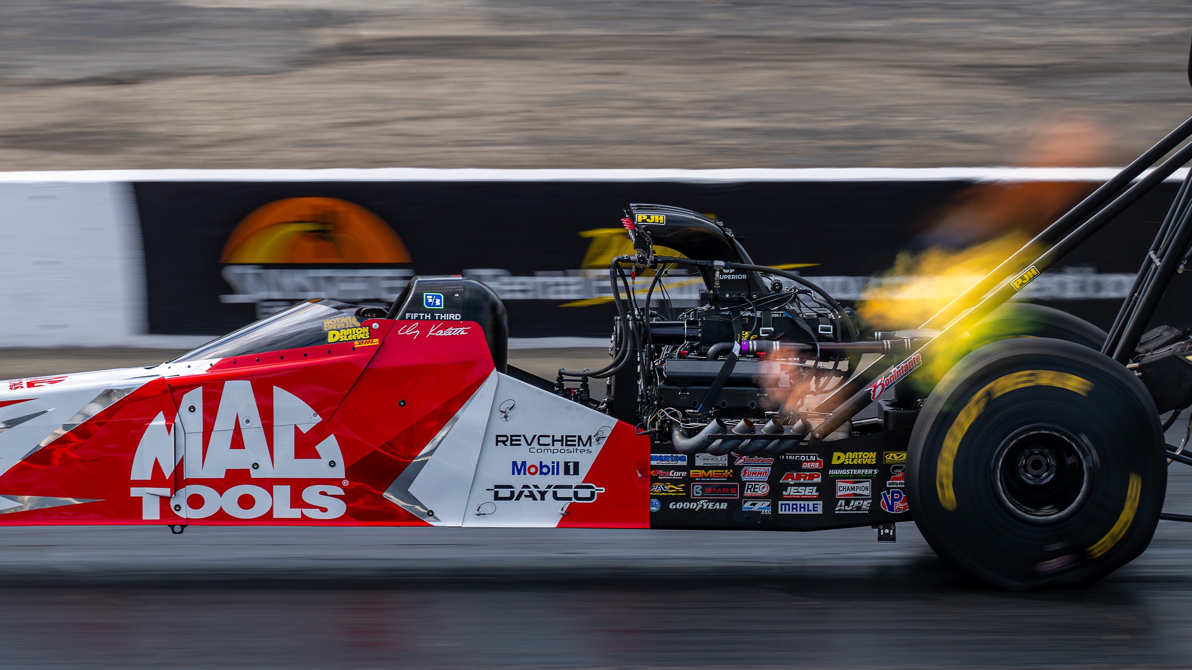 A red and white top fuel dragster panned at speed, header flames trailing, background streaked with motion.