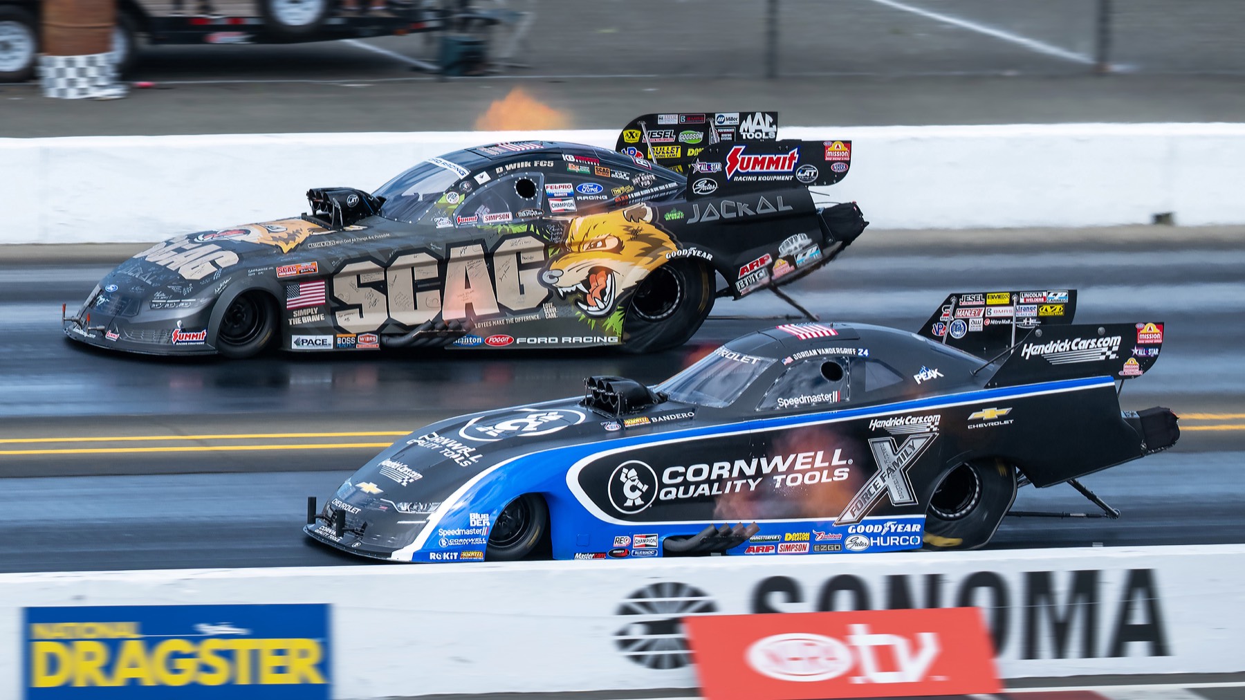 Two funny cars side by side just after launch, seen head-on down the track wall.