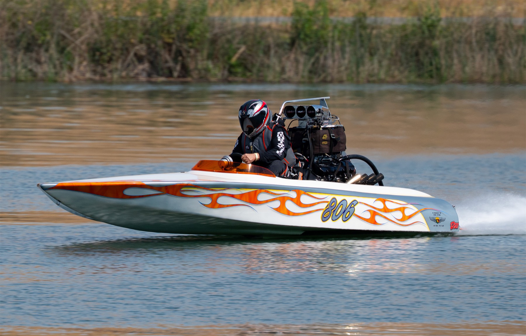 A white drag boat with orange flame paint running hard across calm water.