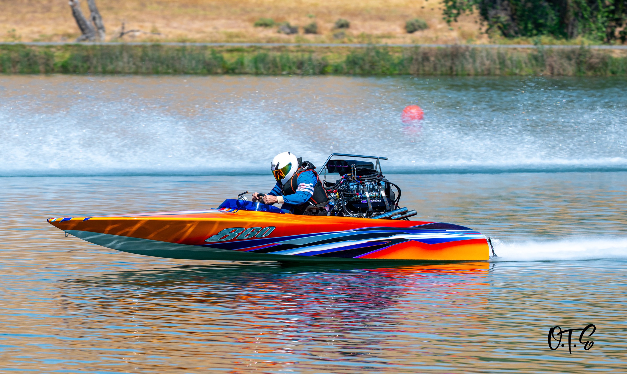 A multicolored drag boat at speed on a lake, throwing a huge rooster tail of spray.